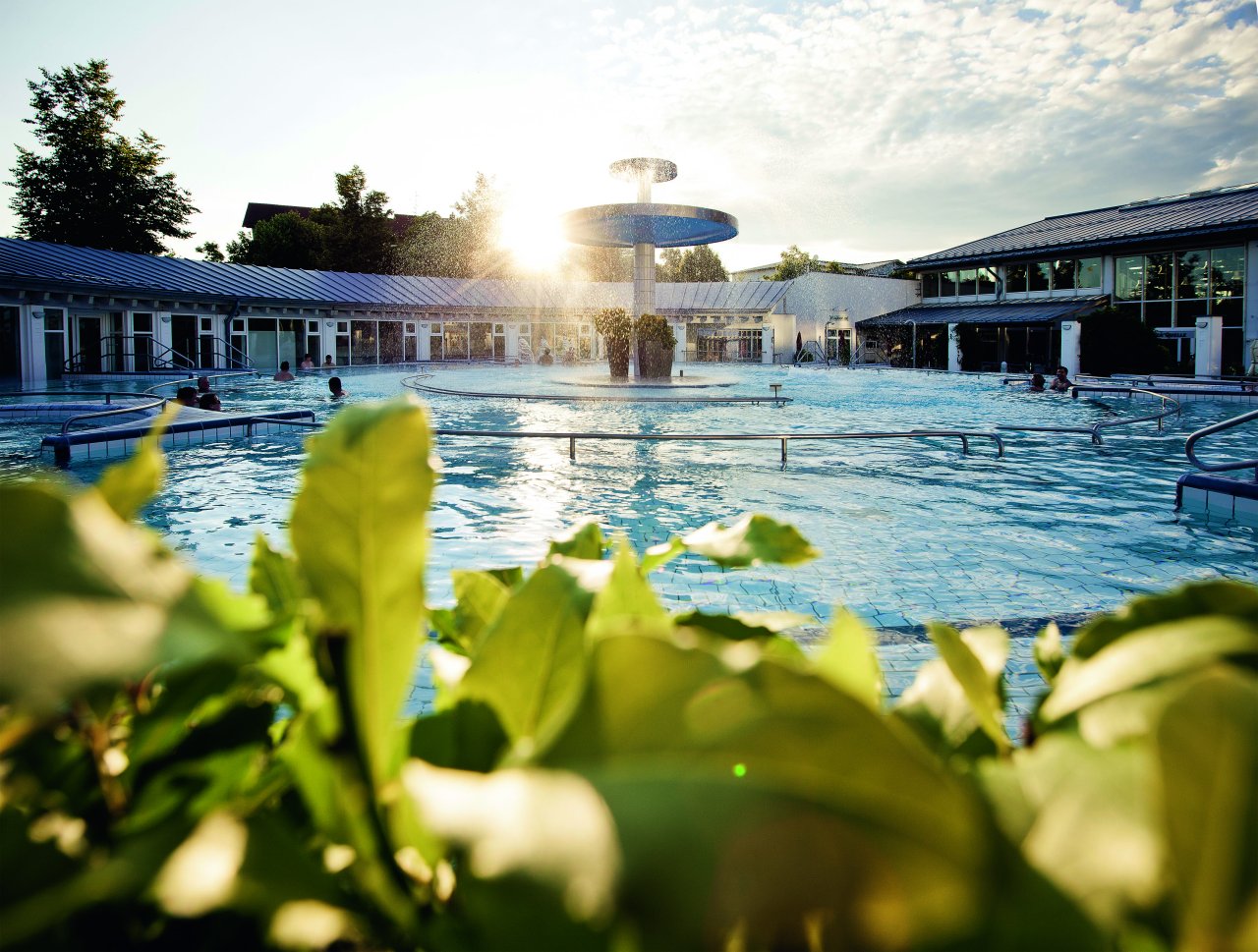 Ein Außenbereich eines Schwimmbads mit einer großen Fontäne in der Mitte. Menschen genießen das Wasser, während die Sonne aufgeht und eine entspannende Atmosphäre schafft. Üppiges Grün umrahmt den Poolbereich.