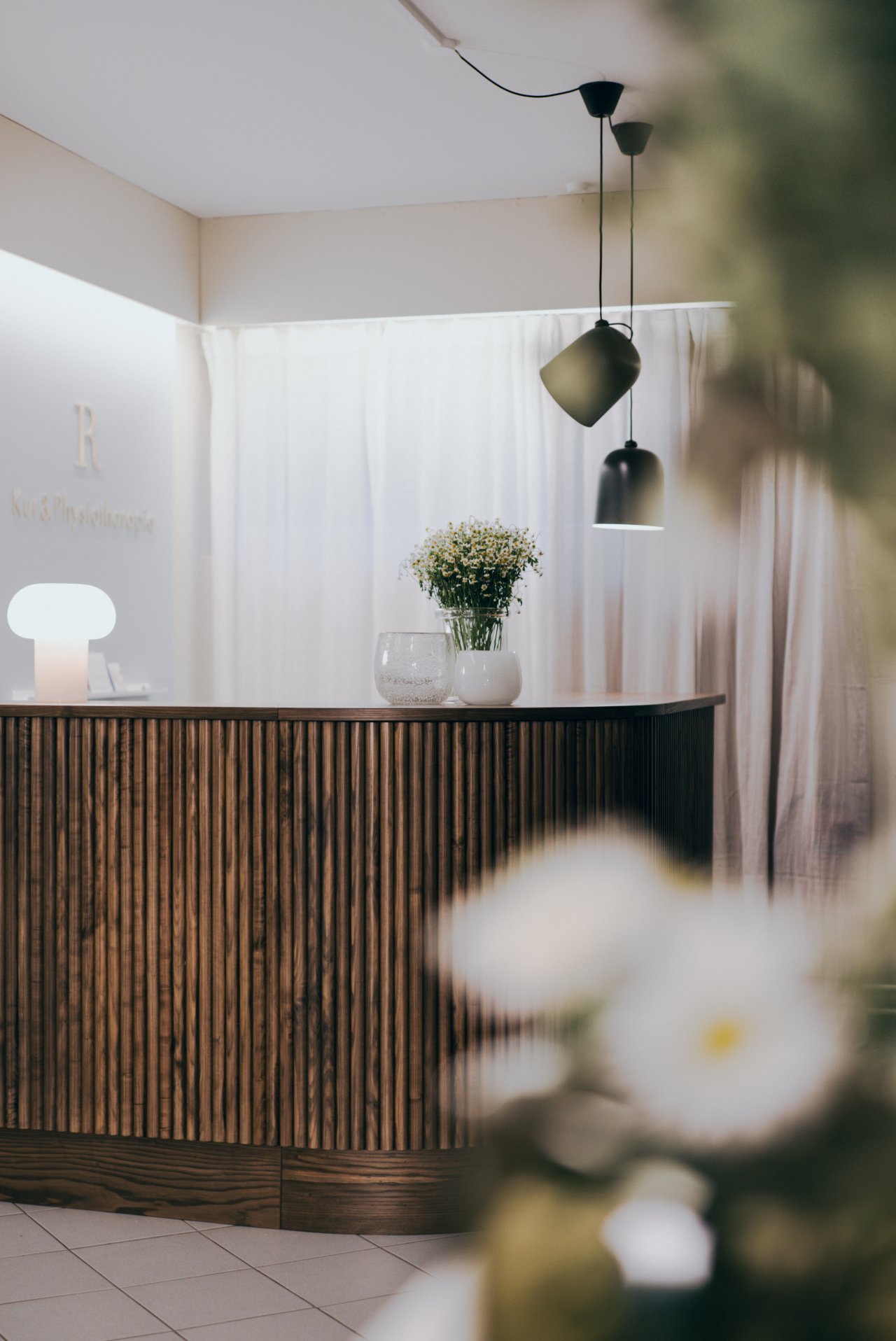 Ein moderner Empfangsbereich mit einem Holztheken, einem hellen Tischlicht und einer blühenden Blumenvase. Dezente Lampen hängen von der Decke. Der Raum hat eine elegante und einladende Atmosphäre.