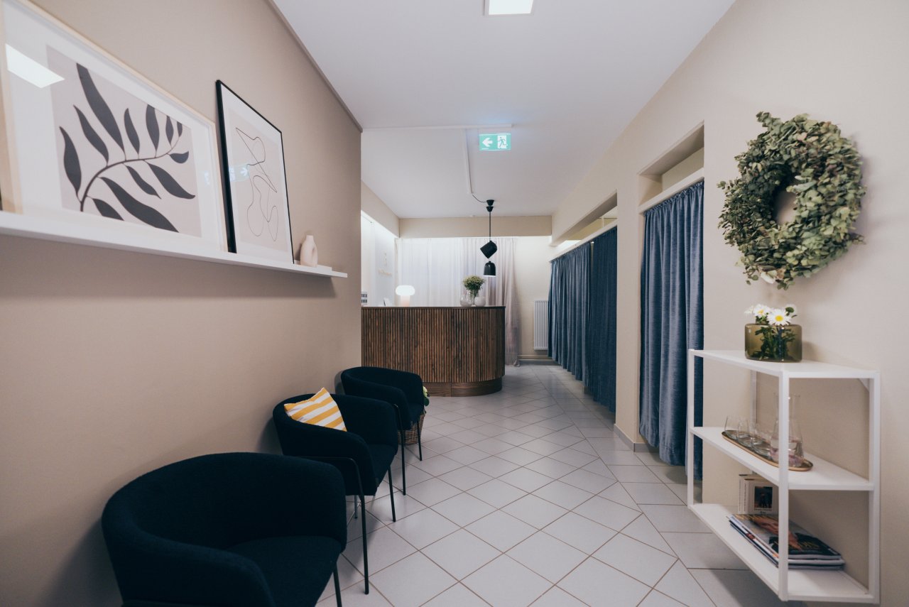 Ein helles Foyer mit grau-blauen Sesseln, einer Holztheke im Hintergrund und einer dekorativen Wand mit Bildern und einem Kranz aus Pflanzen. Vorhänge und eine offene Regale sind ebenfalls sichtbar.