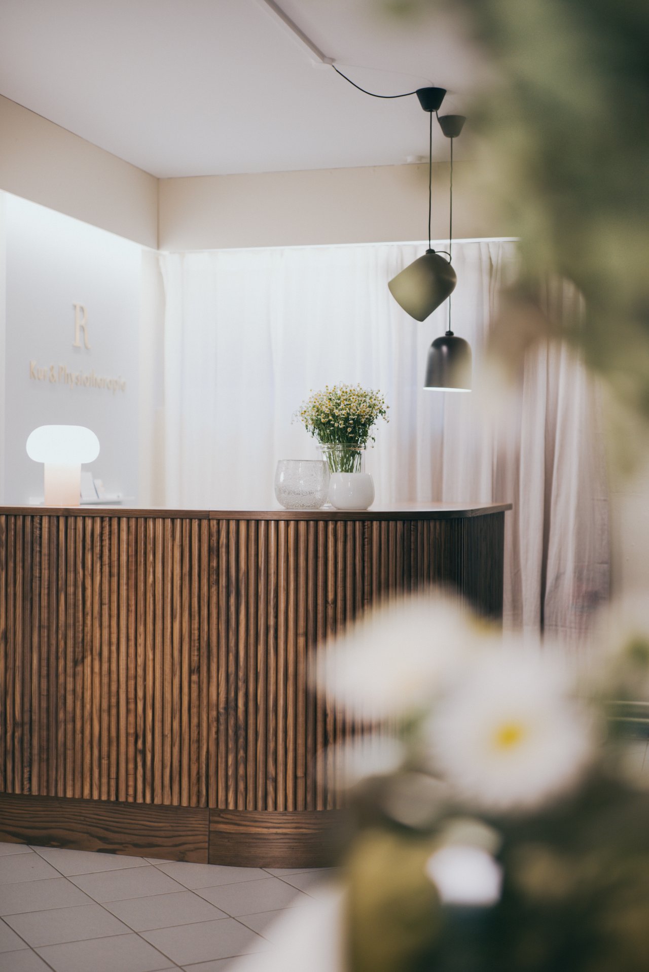 Ein Empfangstresen aus Holz mit einer weißen Lampe und einer Blumenvase. Im Hintergrund hängen Vorhänge und es sind moderne Lampen sichtbar.